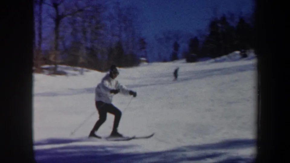 1962 Vermont home-movie frame of a skier crossing a snowy slope, with distant skiers and winter trees behind.