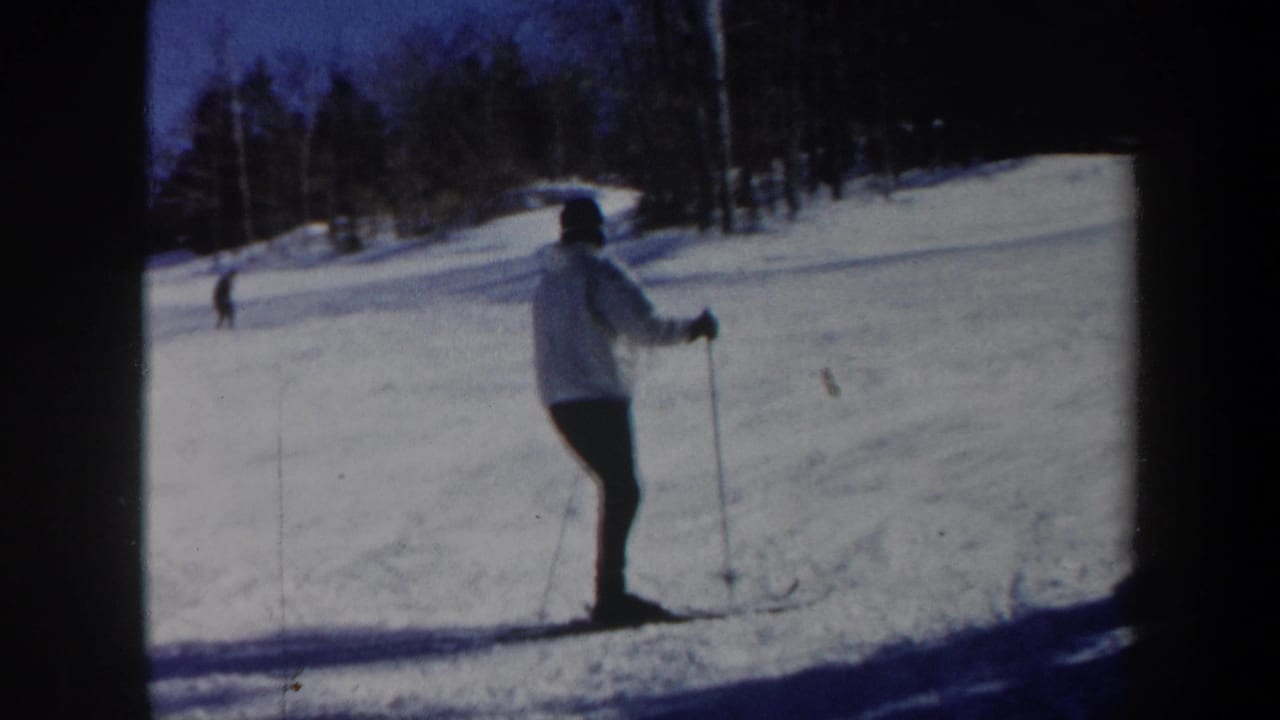 Skier stands or glides near the right side of a snowy run bordered by trees.