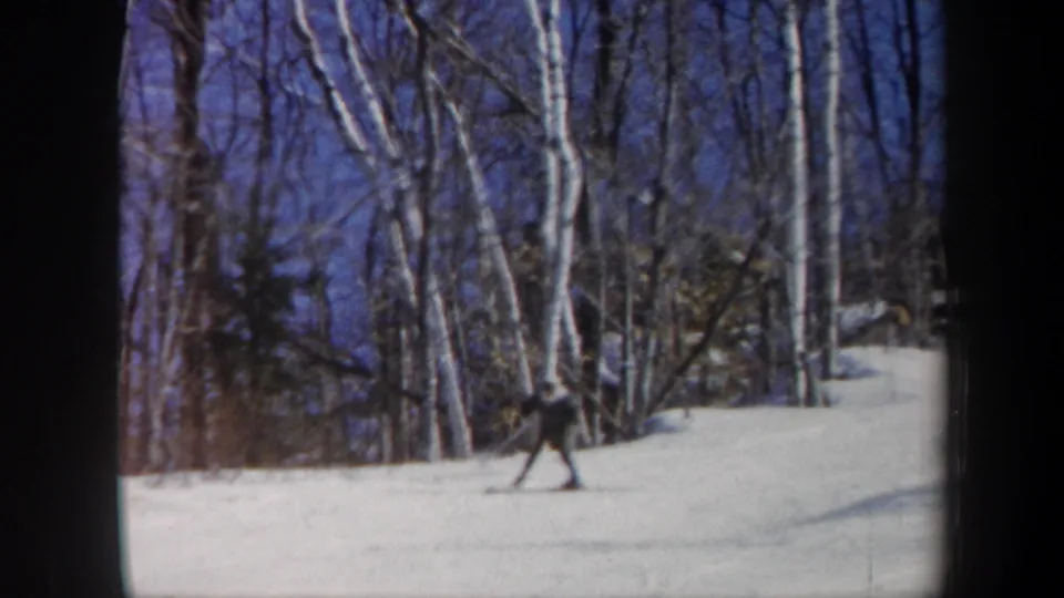 1962 Vermont 8mm frame showing a skier in dark clothing moving along the lower part of a snowy hill near birch trees.