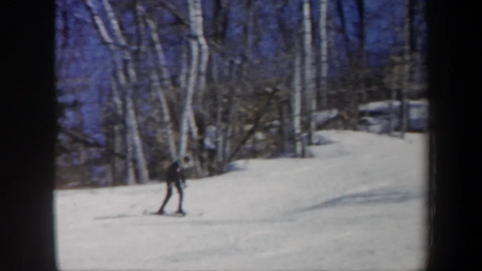 1962 Vermont home-movie frame with the skier continuing across the snow near bare birches and a gentle slope rise.