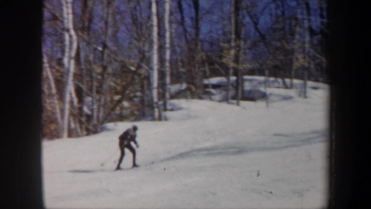 1962 Vermont home-movie frame showing a skier in dark winter clothing moving across a gentle snowy slope with bare trees behind.