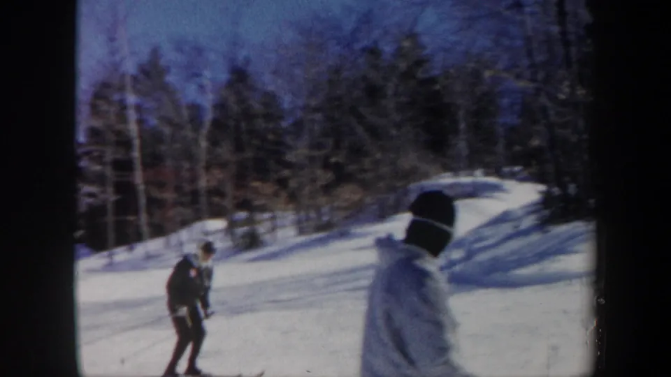 Final 1962 Vermont frame with a bundled observer in the foreground and a dark-clothed skier on the snowy wooded slope beyond.
