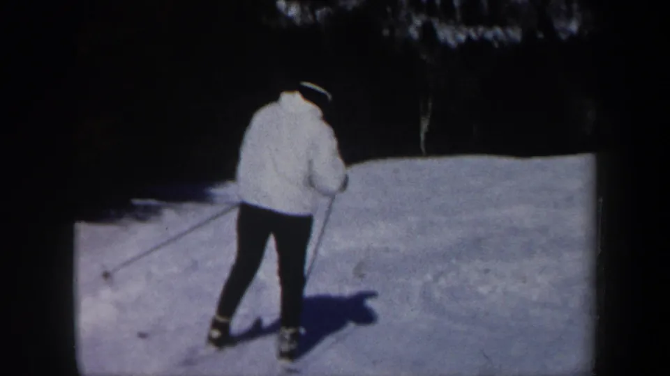 A skier in a white jacket and dark hat is filmed from behind while using poles on a snowy Vermont slope.