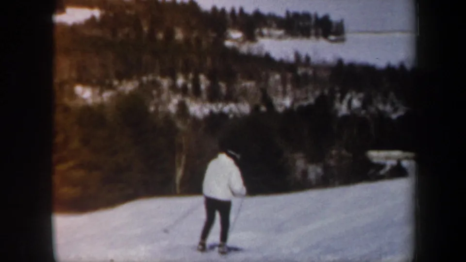 1962 Vermont home-movie frame of a skier in white on a slope overlooking wooded hills and a distant lake or water body.