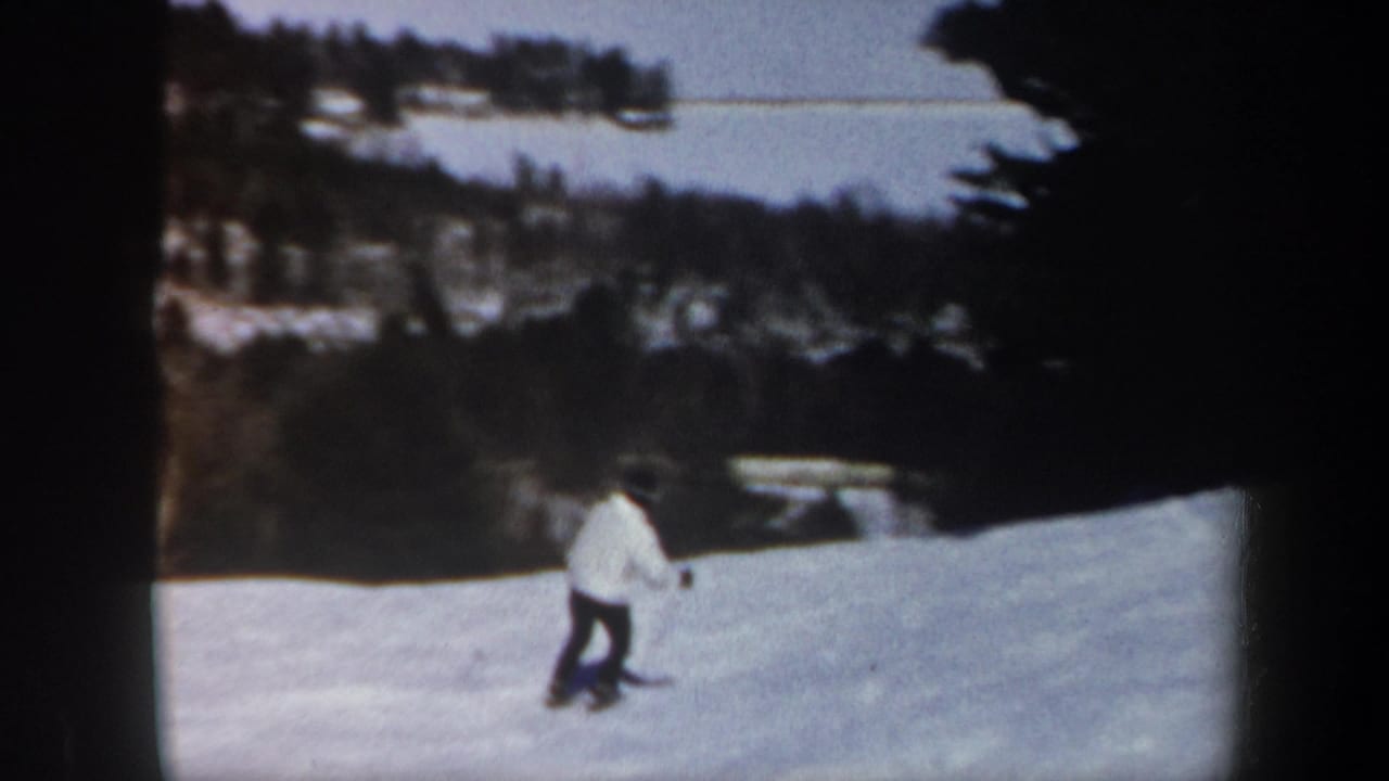 1962 Vermont home-movie frame showing a skier in a white jacket descending a snowy slope bordered by trees and distant hills.