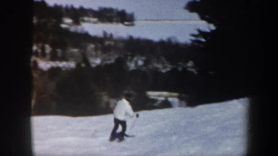 1962 Vermont home-movie frame showing a skier in a white jacket descending a snowy slope bordered by trees and distant hills.