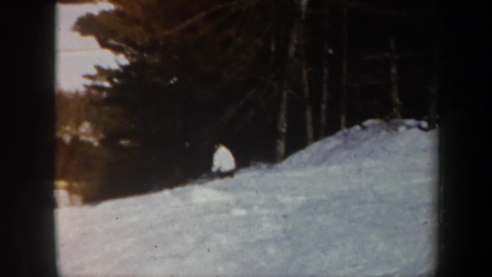 1962 Vermont home-movie frame showing the skier low on the snowy slope near a stand of trees in soft, grainy color.