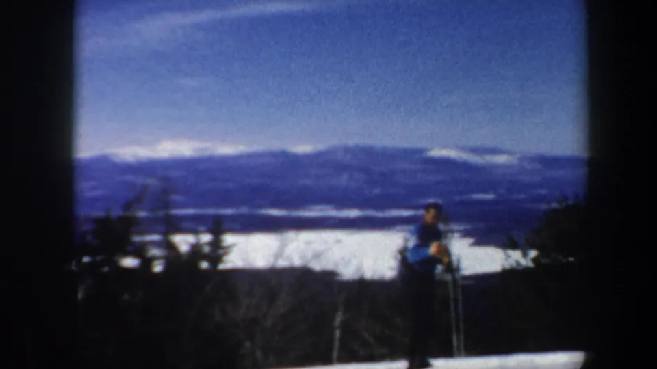 1962 Vermont home-movie frame with a skier paused near trees on snow, backed by a broad mountain range under blue sky.