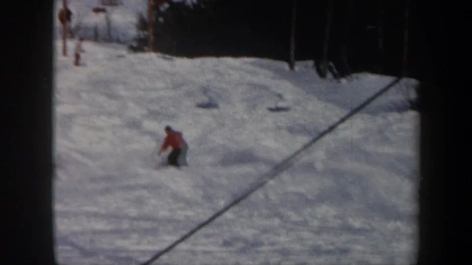 Vermont 1962 home-movie frame showing a red-jacketed skier crouched while descending a snow-covered slope bordered by trees.