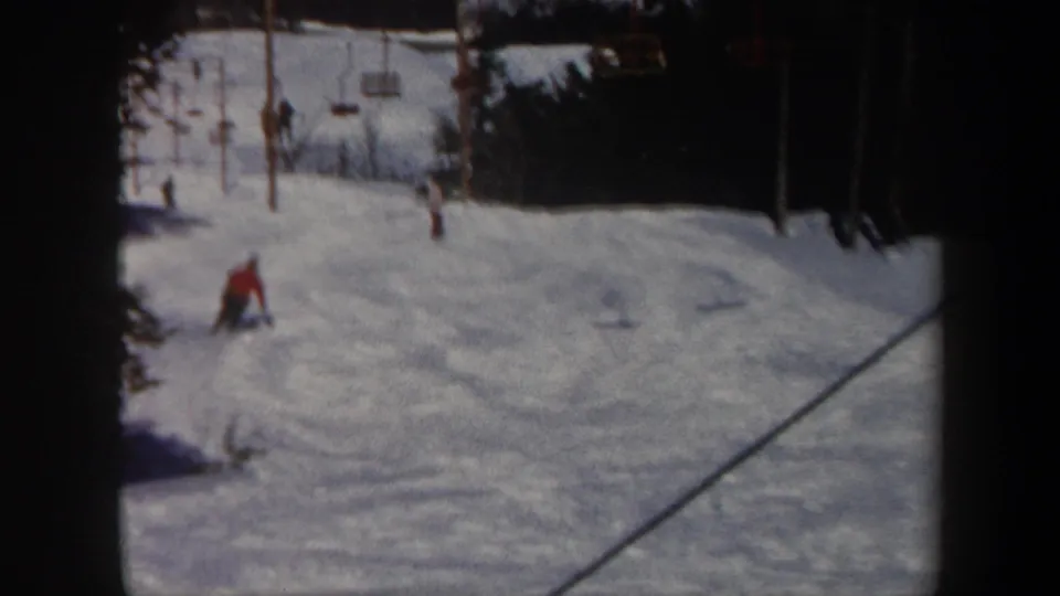 Vermont 1962 frame showing the skier farther down the slope, with trees, lift equipment, and cable lines visible around the ski run.
