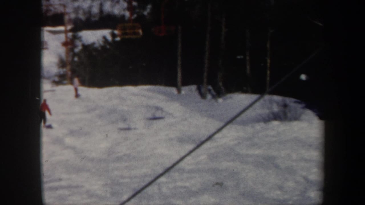 1962 Vermont home-movie frame showing a snowy ski slope, distant skiers, yellow chairlift seats, and trees beyond the run.