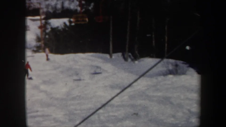 1962 Vermont home-movie frame showing a snowy ski slope, distant skiers, yellow chairlift seats, and trees beyond the run.