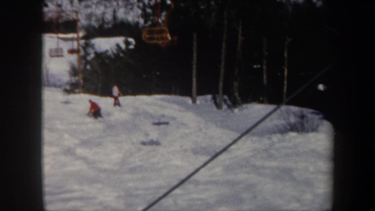 Small figures ski on a snowy slope with yellow chairlift seats overhead and trees behind.