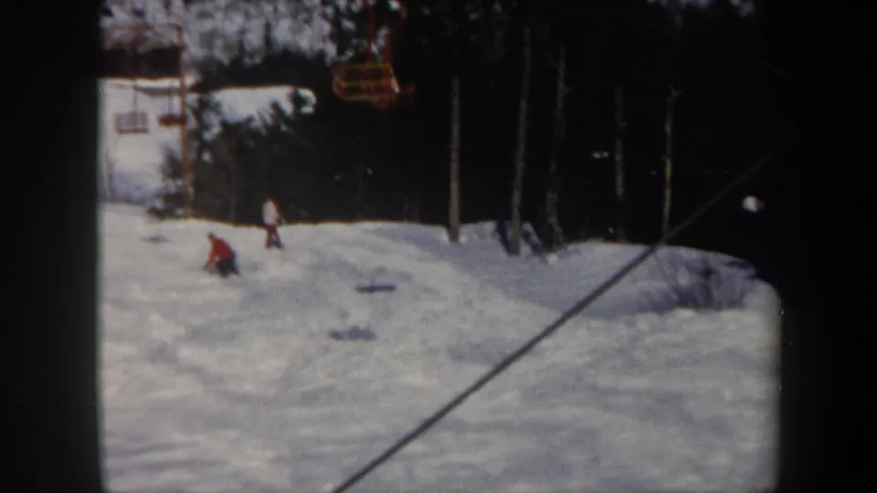 1962 Vermont ski area view with several distant skiers on the snow, yellow lift chairs above, and a tree line behind the run.