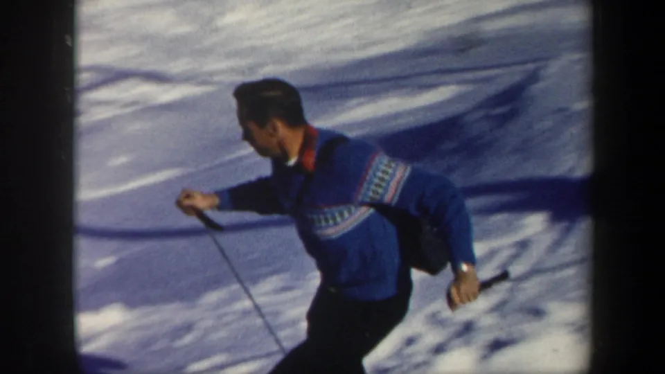 1962 Vermont home-movie frame of an adult skier in a blue patterned sweater carrying poles across the snow.