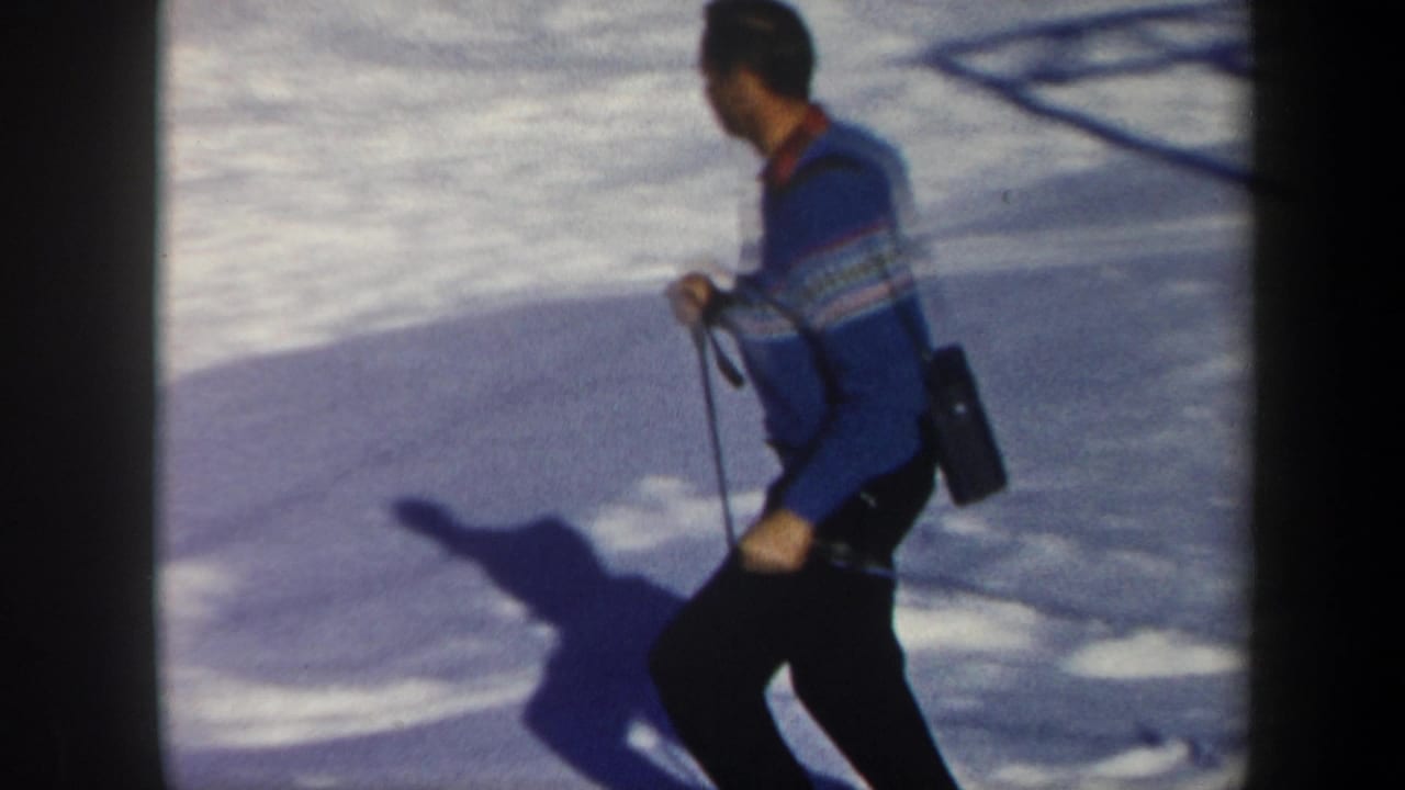 In 1962 Vermont home-movie footage, an adult skier in a blue patterned sweater moves across snow with ski poles and a shoulder bag.