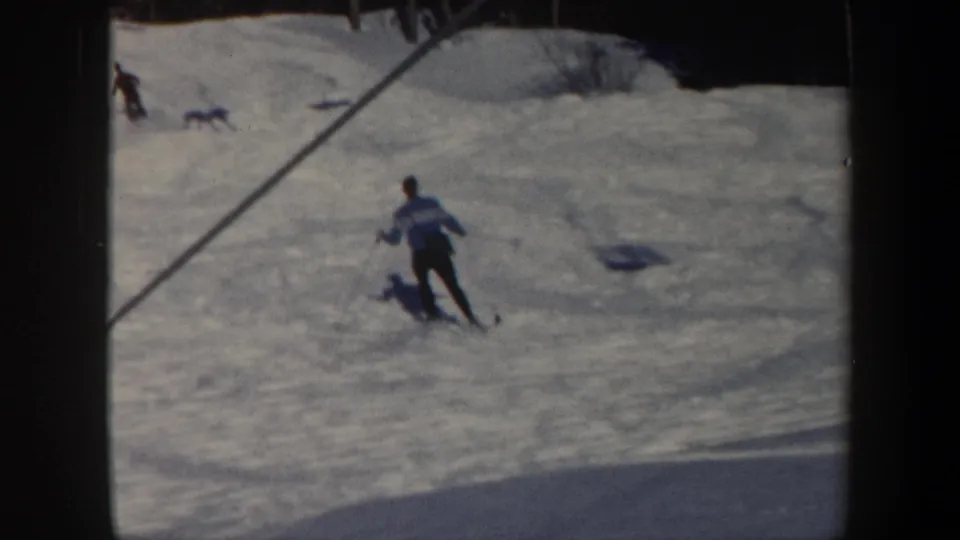 1962 Vermont 8mm frame showing a skier in a light and blue jacket moving downhill on a snowy slope with other skiers in the distance.