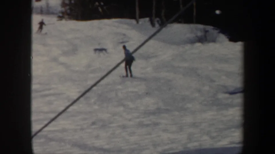 1962 Vermont ski-slope frame with a skier near a diagonal tow or lift line, distant skiers, and bright snow under natural light.