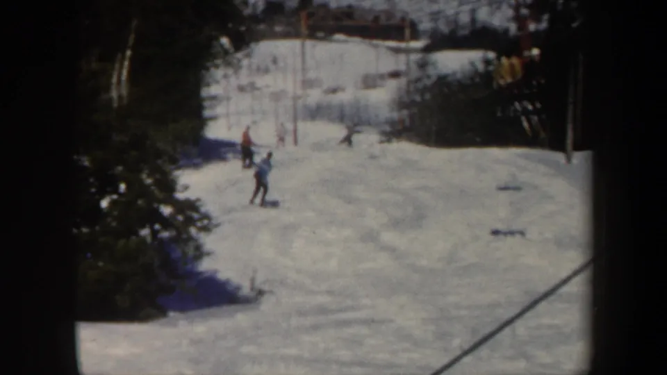 1962 Vermont 8mm frame with several skiers spread across a snowy slope, evergreen trees at left, and a diagonal lift or tow line crossing the view.
