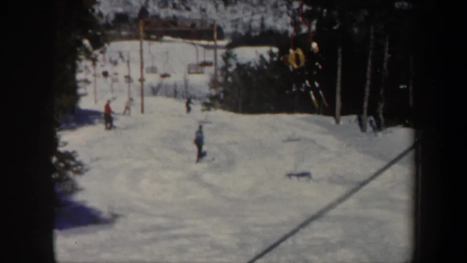 1962 Vermont 8mm frame showing skiers on a snowy slope near chairlift towers and dark evergreen trees.