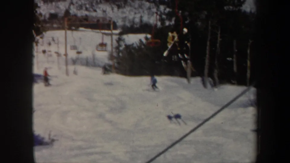1962 Vermont ski slope with distant skiers, red chairlift chairs, evergreens, and soft 8mm film grain.