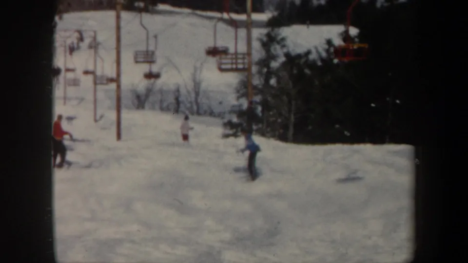 1962 Vermont home-movie frame of skiers near red chairlift chairs and lift poles on a snow-covered slope.
