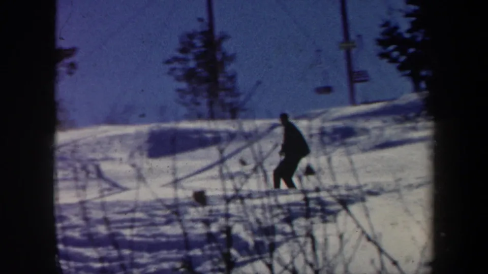 1962 Vermont 8mm frame with a dark-clad figure crossing a snowy ski area near chairlift towers and winter trees.
