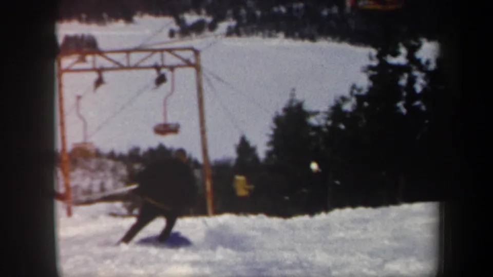 A skier moves downhill near a ski-lift frame, suspended lift seats, snow, and evergreen trees in 1962 Vermont footage.