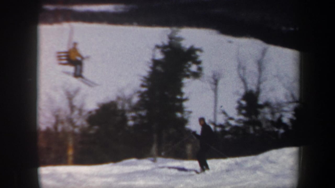 1962 Vermont home-movie frame showing a skier on snow while chairlift riders pass above the slope.