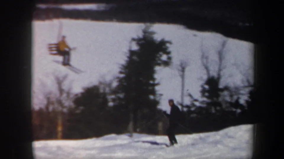 1962 Vermont home-movie frame showing a skier on snow while chairlift riders pass above the slope.