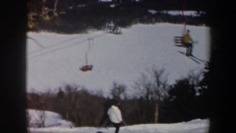 1962 Vermont ski-resort frame with a white-jacketed skier below chairlift seats and winter tree lines.