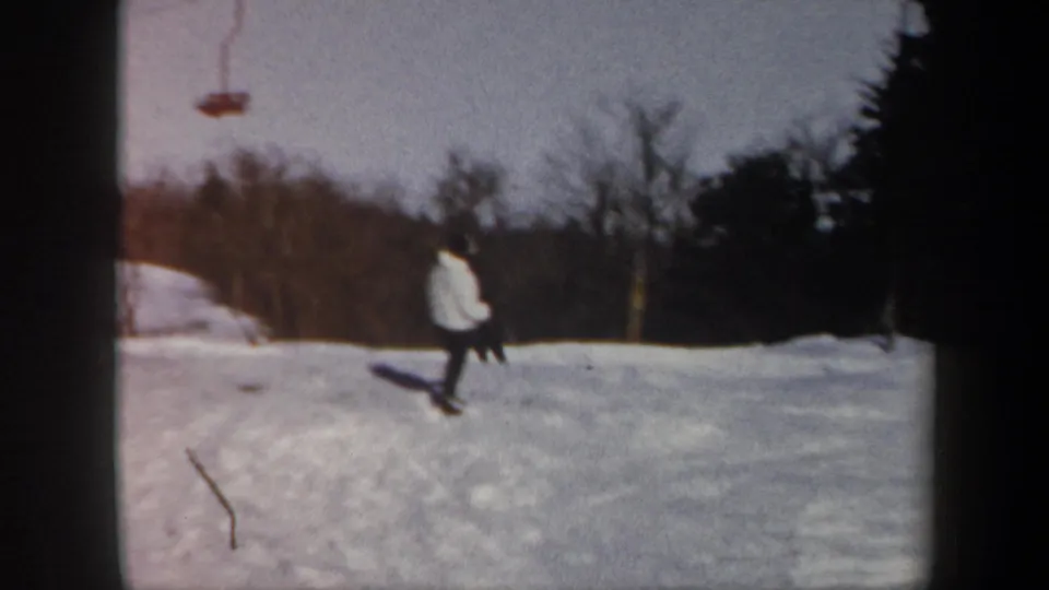1962 Vermont home-movie frame of a white-jacketed skier on a snowy resort slope near bare trees and lift equipment.