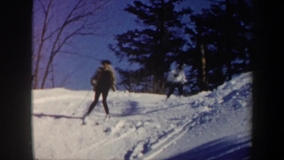 1962 Vermont frame with two young people skiing on a bright winter slope, framed by bare trees and evergreens.