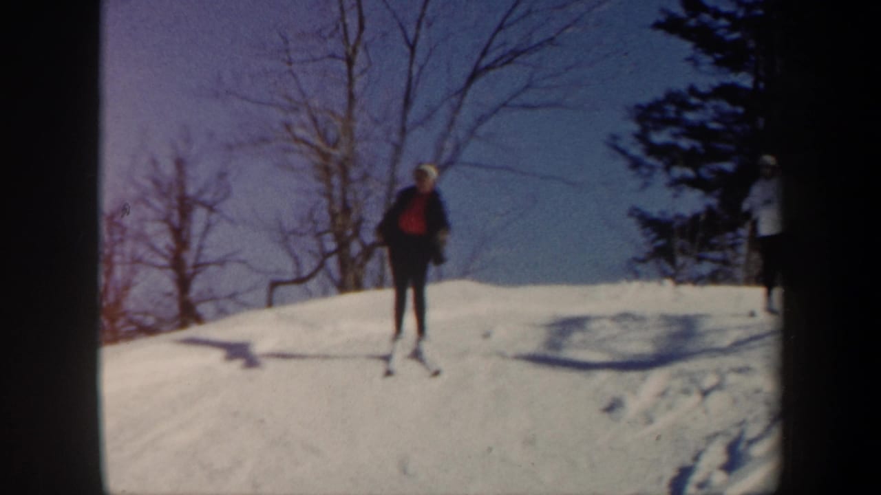 1962 Vermont 8mm frame of a skier in a dark jacket and red top moving across a snowy hillside with winter trees behind.