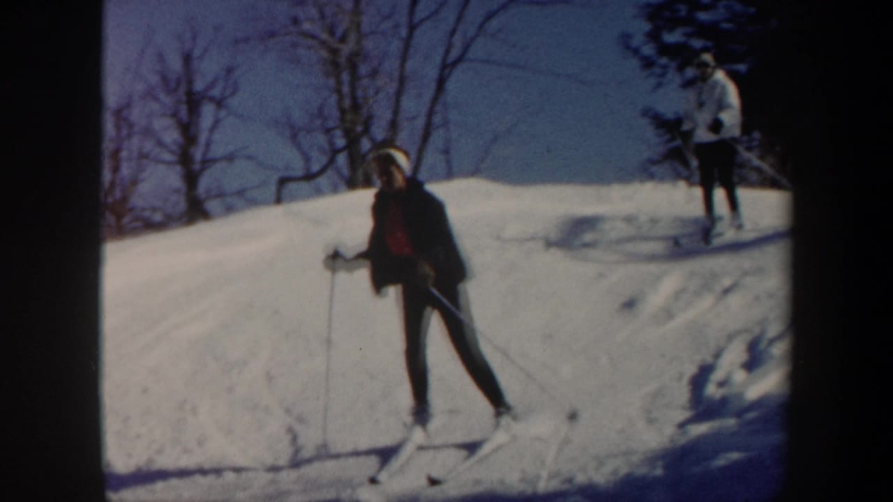 Skier leaning on poles on a snowy hill while another skier stands in the background.