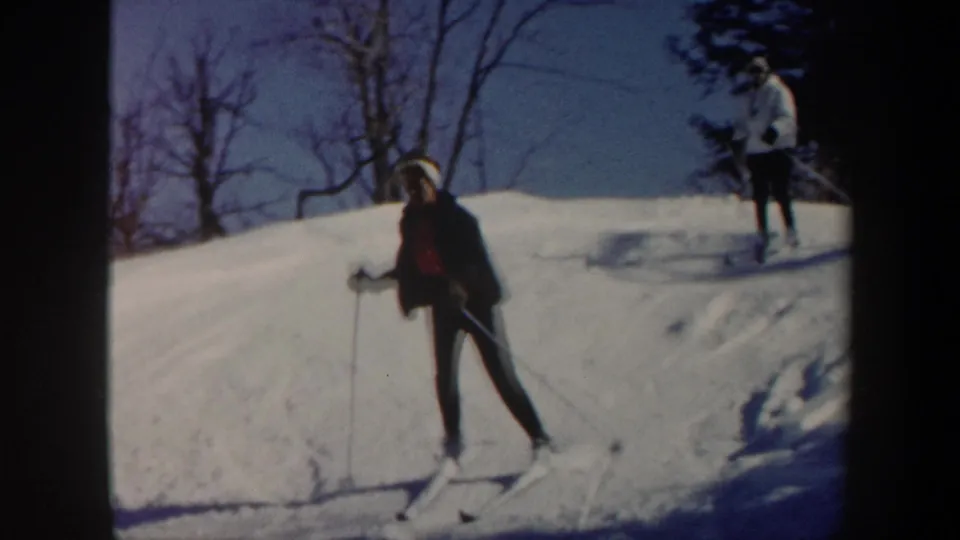 1962 Vermont home-movie frame showing a skier on a snow-covered slope with a second skier in white near the upper right.