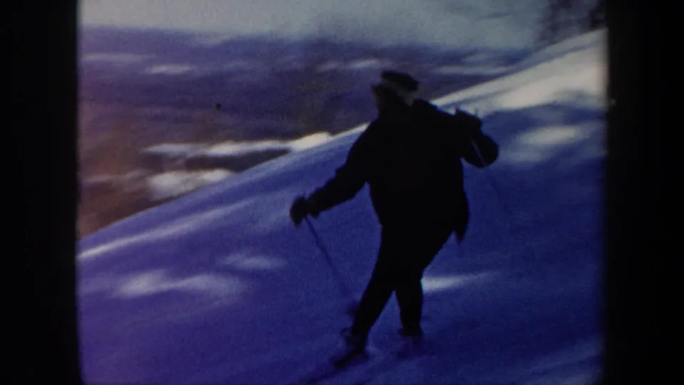 1962 Vermont 8mm frame with a dark, partly silhouetted skier moving across a shaded snow slope near a distant landscape view.