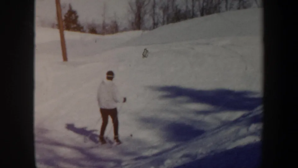 1962 Vermont home-movie frame of a skier in a light jacket on a snowy slope, with bare trees and blue shadows visible.