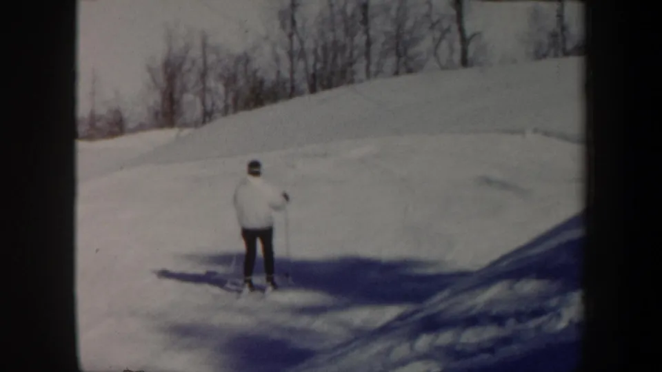 1962 Vermont 8mm frame showing a distant skier in a white jacket and dark pants on a snowy hillside lined with bare trees.