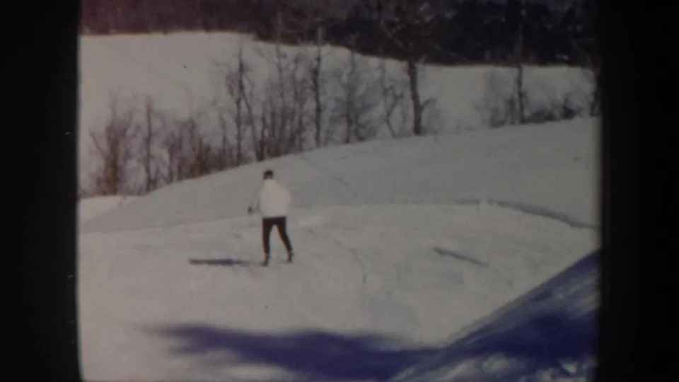 1962 Vermont 8mm frame with a lone skier crossing a bright snow slope below a line of bare winter trees.