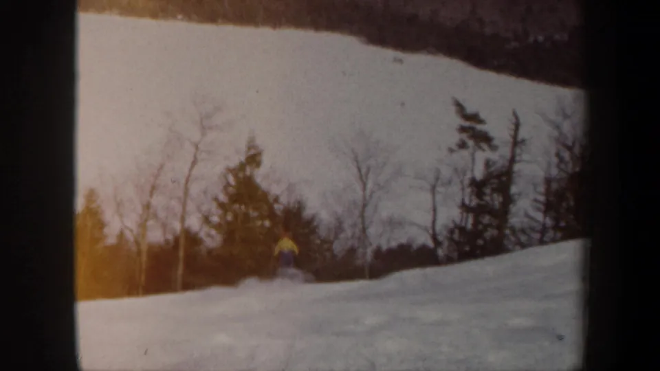 1962 Vermont 8mm frame showing a distant skier in a yellow jacket on a snow-covered hill below bare trees and evergreens.