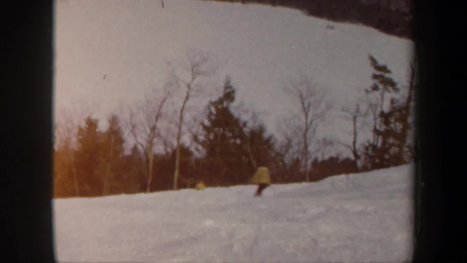 1962 Vermont home-movie frame with small yellow-jacketed skiers crossing a white slope against a winter tree line.