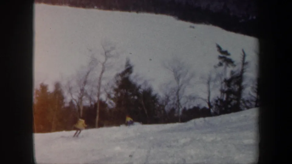 1962 Vermont 8mm frame showing one skier in yellow nearer the camera and another small skier farther across the snowy hill.