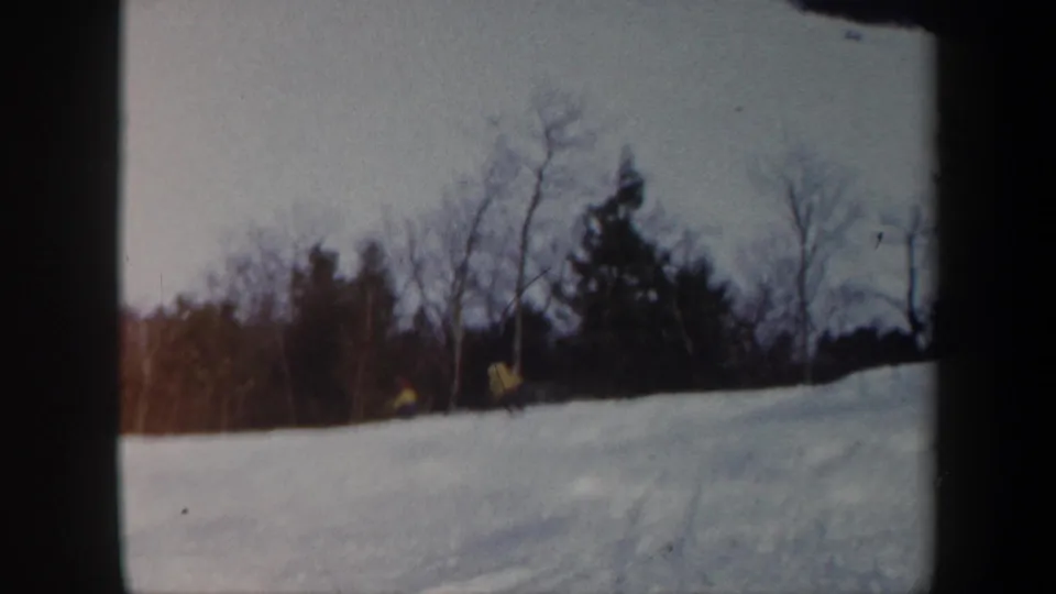 1962 Vermont home-movie frame with two small skiers in yellow jackets on a snowy slope below bare trees and evergreens.