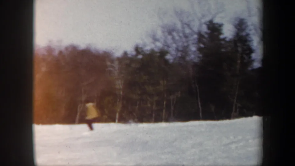 A skier in a light-colored jacket descends a Vermont snow slope with dark evergreens and leafless trees behind.