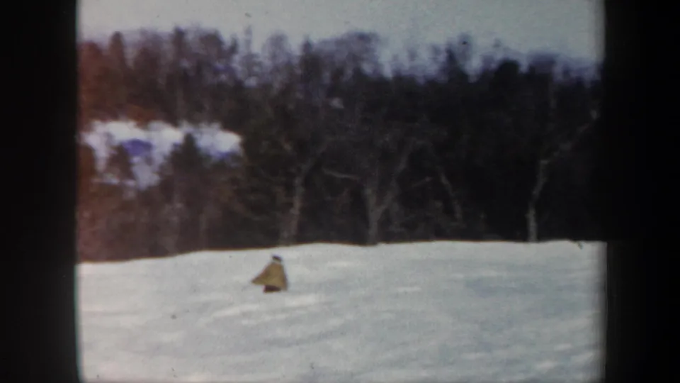 Home-movie frame of a skier descending lower across a snowy Vermont hillside beneath a line of winter trees.