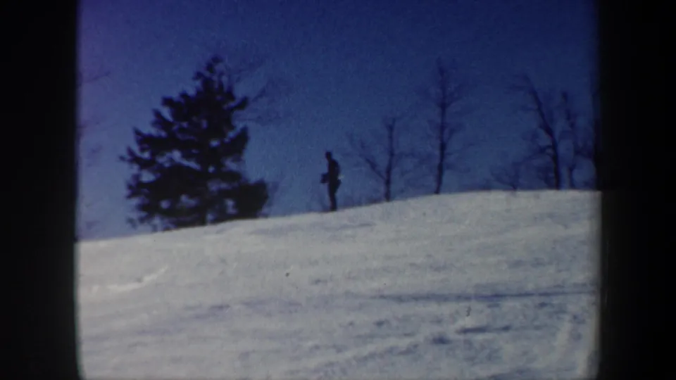 1962 Vermont 8mm frame showing a lone skier crossing a snowy hill beside an evergreen and leafless trees.