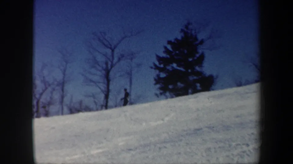 1962 Vermont home-movie frame with a distant skier low on the hill, an evergreen, bare trees, and blue-toned snow.