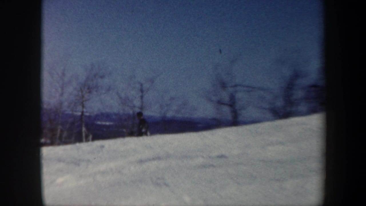 A distant skier appears near the crest of a snowy Vermont hillside, framed by bare trees and blue sky.