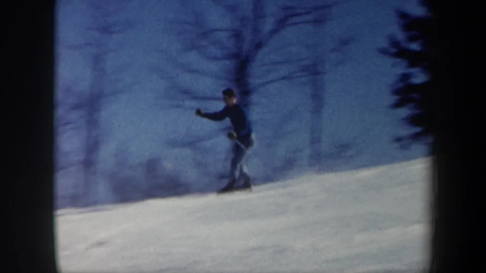 A lone skier crosses the snow-covered hillside in 1962 Vermont, with bare trees and blue sky behind.